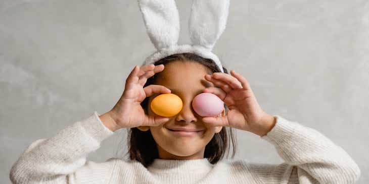 Uma pessoa usando orelhas de coelho lúdicas e segurando pequenos ovos de páscoa pintados no lugar dos olhos.