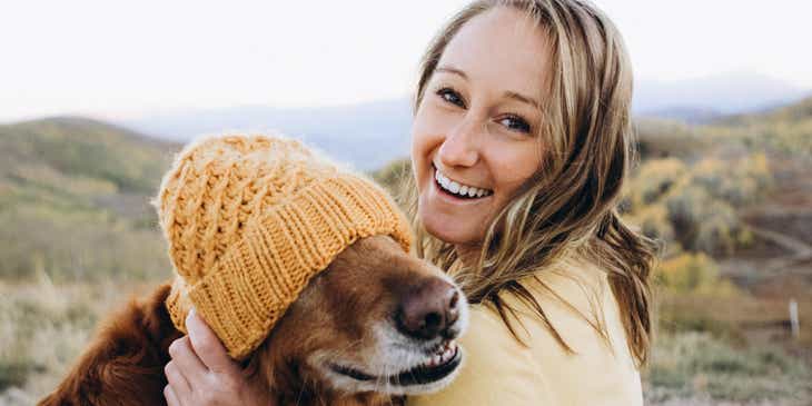 A person with a pleasant smile holding a dog that is wearing a beanie.