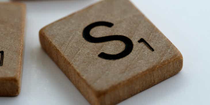 Ein rechteckiger Scrabble-Holzstein mit dem Buchstaben „S“ und einer kleinen Ziffer „1“ liegt neben zwei ähnlichen Spielsteinen auf einer weißen Oberfläche.