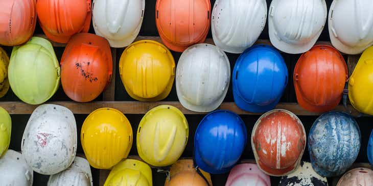 Verschiedenfarbige Helme, die an Mitarbeiter als Schutzmaßnahme im Rahmen von Arbeitssicherheitsbestimmungen ausgegeben werden, hängen an einem Holzregal.