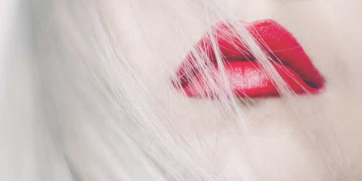 Das Gesicht einer Frau in sexy Pose, deren platinblonde, lange Haaren locker über die rot geschminkten Lippen fallen.