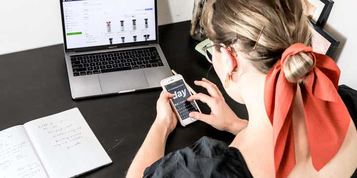 Una mujer con su teléfono en la mano y frente a su computadora en una agencia de Social Media.