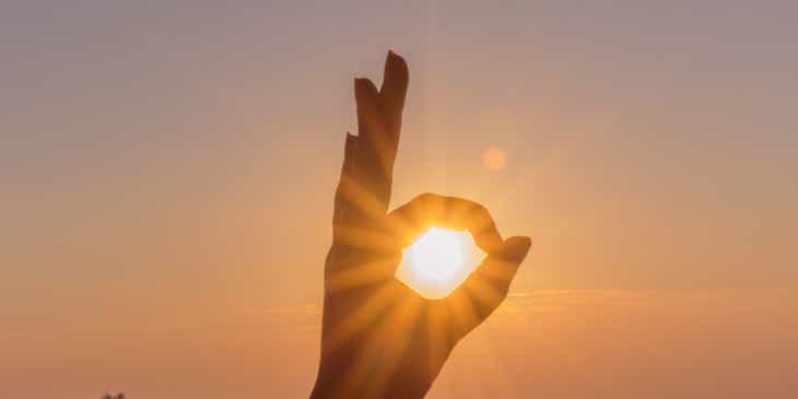 Zbliżenie na zachodzące słońce ujęte w środku dłoni wykonującej palcami znak „ok”.