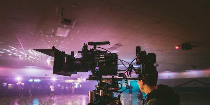 Ein Mitarbeiter einer Fernsehproduktionsfirma steht neben einer Kamera in einer erleuchteten Halle und filmt eine Szene.