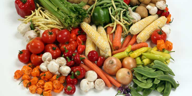 Una pila de distintas verduras, como zanahorias, apio, maíz y cebollas, sobre fondo blanco.