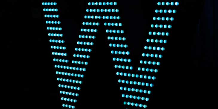 Eine hellblau leuchtende LED-Buchstabenlampe in der Form eines "W" vor einem dunklen Hintergrund.